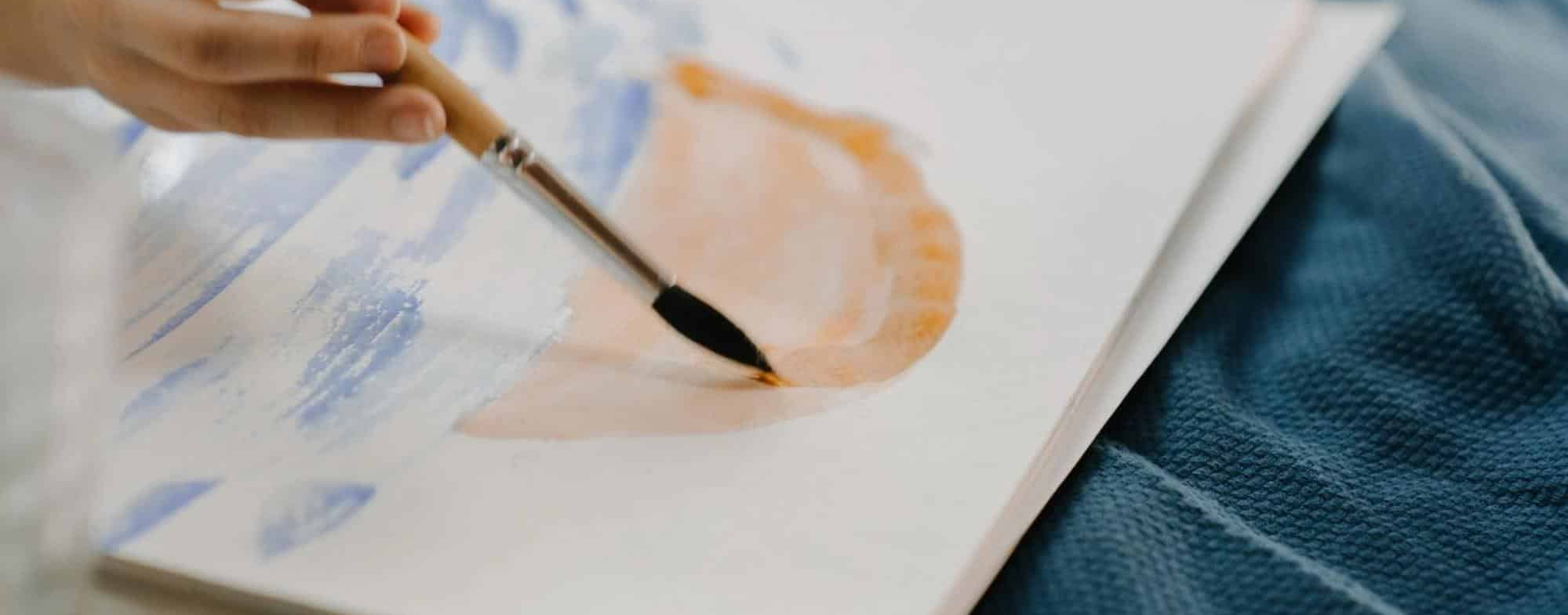 Close-Up Of Paintbrush Applying Soft Orange Watercolor On Paper With Blue Strokes, Artistic Creative Process_1