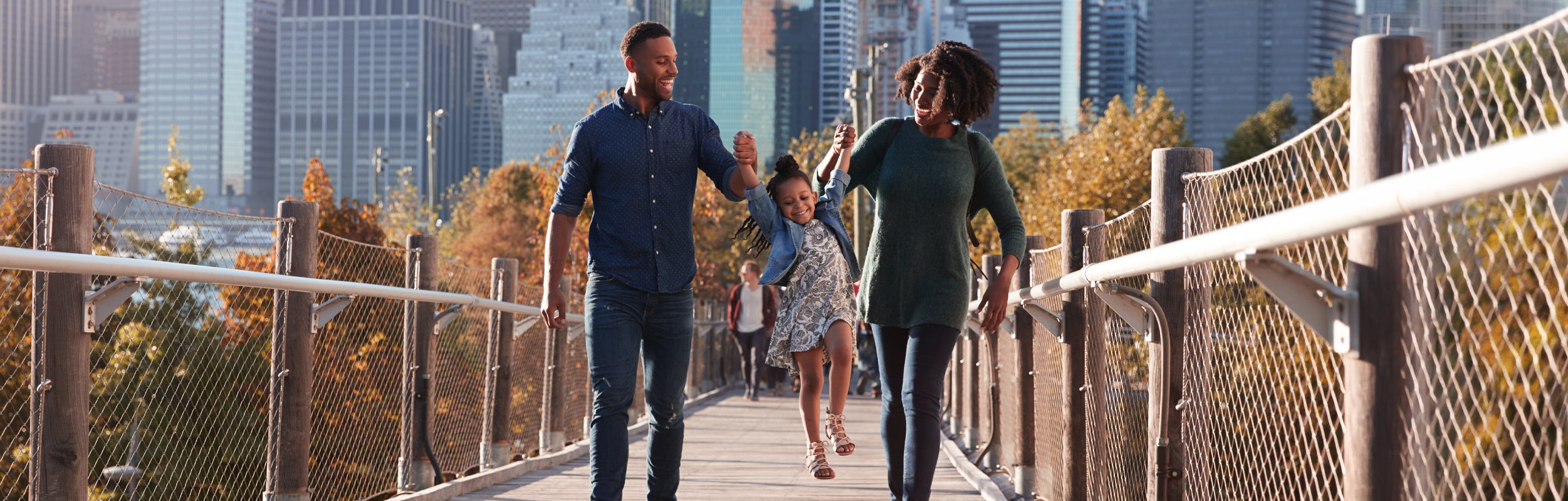 Family Walking Across Urban Park Boardwalk, Child Lifted Midstride, Autumn Foliage And City Skyline Backdrop
